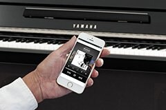 A person holding a white smartphone displaying an audio player app in front of a black Yamaha digital piano with visible white and black keys.