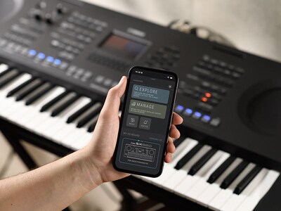 PSR-SX620 and a hand holding a smartphone with the Yamaha official app “Expansion Explorer” screen.