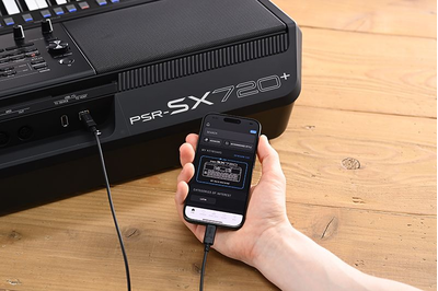 Yamaha Arranger Workstation PSR-SX720+ and a hand holding smartphone with the Yamaha official app “Expansion Explorer” screen.