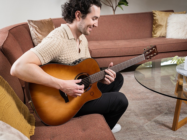 A smiling man sits on a couch in a stylish living room playing the TAG3 C guitar in Sand Burst finish.